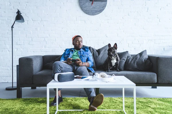 Digitale Geräte auf dem Tisch vor einem jungen Mann, der einen Steuerknüppel hält und auf einem Sofa bei einer französischen Bulldogge sitzt — Stockfoto