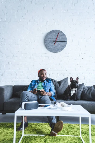 Young man holding joystick while sitting on sofa with bulldog and digital gadgets on table — Stock Photo