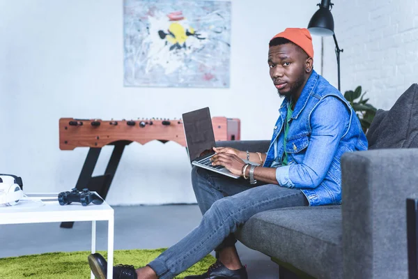 Bello afro-americano che lavora sul computer portatile mentre è seduto sul divano — Foto stock