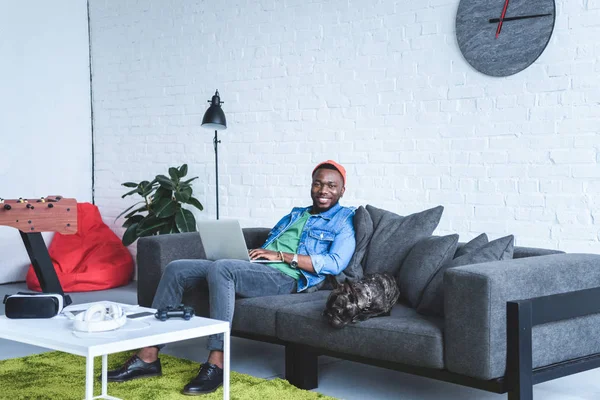 Moderne Gadgets auf dem Tisch vor einem afrikanisch-amerikanischen Mann, der am Laptop arbeitet und auf dem Sofa einer französischen Bulldogge sitzt — Stockfoto