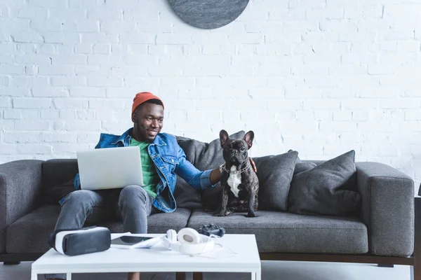 Uomo afroamericano che lavora su laptop e abbraccia bulldog francese da tavolo con gadget moderni — Foto stock