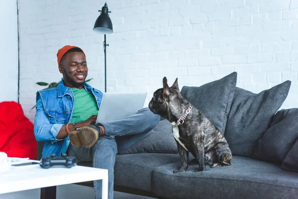 Young man working on laptop while sitting on sofa with bulldog — Stock Photo