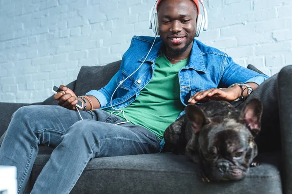 Handsome african american man in headphones listening to music and cuddling French bulldog — Stock Photo