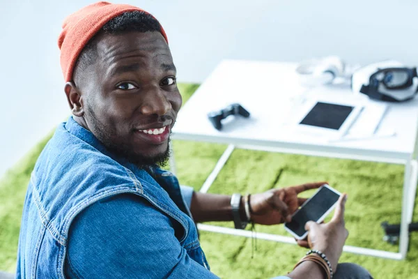 Joven sonriendo y usando smartphone por mesa con gadgets - foto de stock