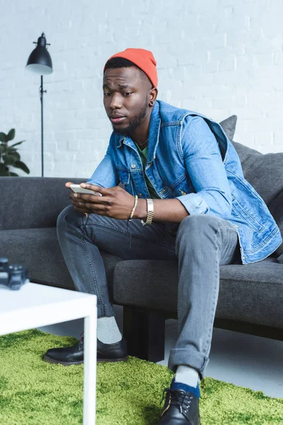 Africano americano homem sentado no sofá e usando smartphone — Fotografia de Stock