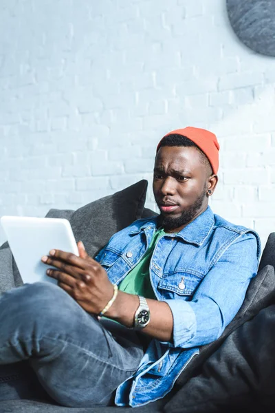 Hombre afroamericano sentado en un sofá y usando una tableta digital - foto de stock