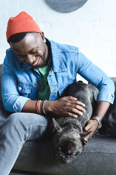 Bel homme afro-américain jouer avec noir Frenchie sur canapé — Photo de stock