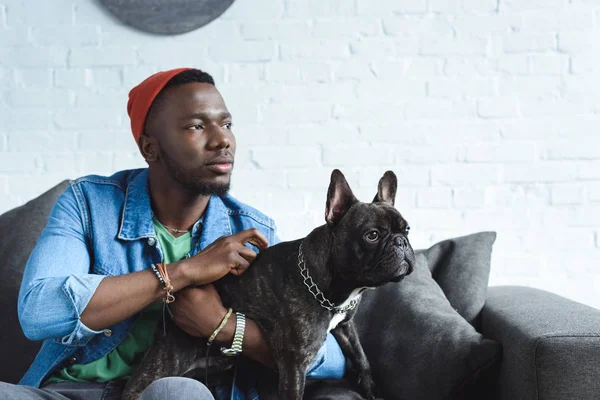 African american man cuddling black Frenchie on sofa — Stock Photo