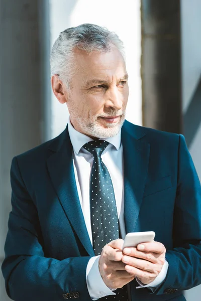 Sonriente hombre de negocios senior sosteniendo el teléfono inteligente y mirando hacia otro lado - foto de stock