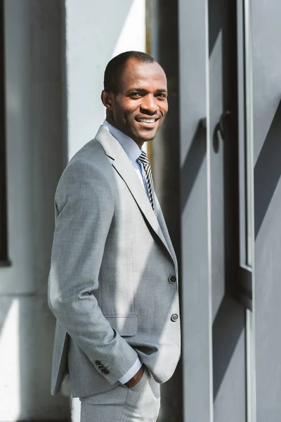Retrato de joven hombre de negocios afroamericano guapo de pie con la mano en el bolsillo y sonriendo a la cámara - foto de stock
