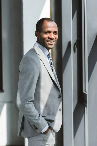 Retrato de belo jovem empresário afro-americano de pé com a mão no bolso e sorrindo para a câmera — Fotografia de Stock