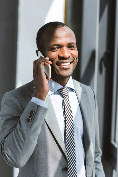 Joven hombre de negocios afroamericano hablando por teléfono inteligente y sonriendo a la cámara - foto de stock