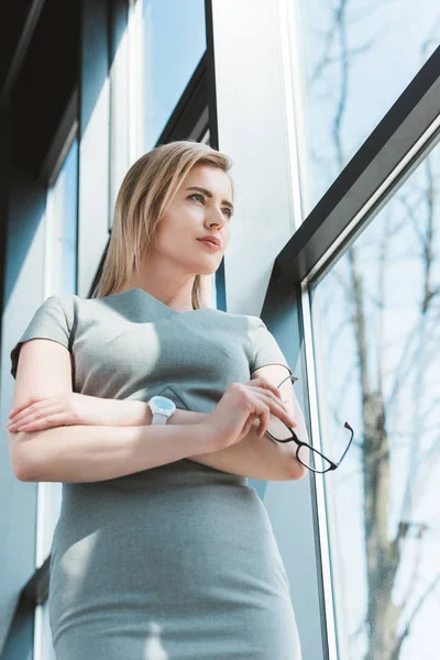 Low angle view of businesswoman standing with crossed arms and holding eyeglasses while standing near window — Stock Photo