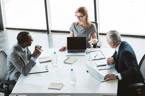 Vista de ángulo alto de la mujer de negocios que muestra el ordenador portátil con pantalla en blanco y mirando a los colegas masculinos durante la reunión - foto de stock