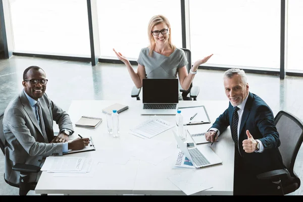 Happy multiethnic business people sitting at table and smiling at camera during successful meeting — Stock Photo
