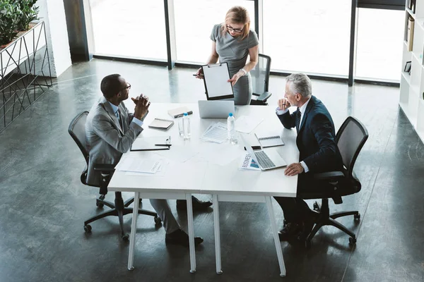 Visão de alto ângulo de empresária mostrando área de transferência em branco para homens de negócios multiétnicos na reunião — Fotografia de Stock