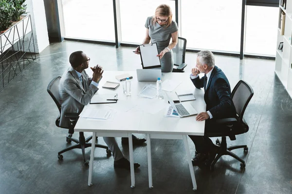 Vue grand angle de femme d'affaires montrant le presse-papiers vierge aux hommes d'affaires multiethniques à la réunion — Photo de stock