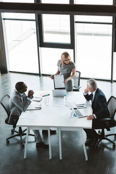 Vista de ángulo alto de la mujer de negocios mostrando portapapeles en blanco a los colegas en la reunión de negocios - foto de stock