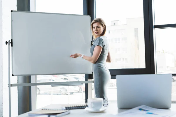 Belle femme d'affaires pointant vers tableau blanc vierge et regardant la caméra — Photo de stock