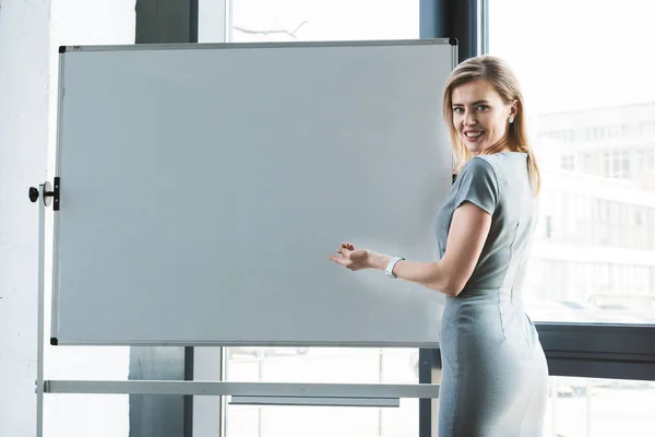 Geschäftsfrau zeigt auf leeres Whiteboard und lächelt in die Kamera — Stockfoto