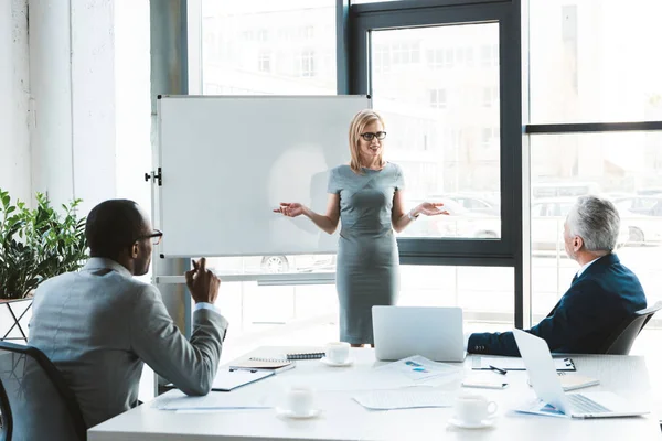 Schöne lächelnde Geschäftsfrau, die neben Whiteboard steht und Kollegen während eines Geschäftstreffens ansieht — Stockfoto