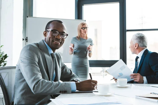 Junger afrikanisch-amerikanischer Geschäftsmann macht sich Notizen und lächelt in die Kamera, während er sich bei einem Geschäftstreffen mit Kollegen unterhält — Stockfoto