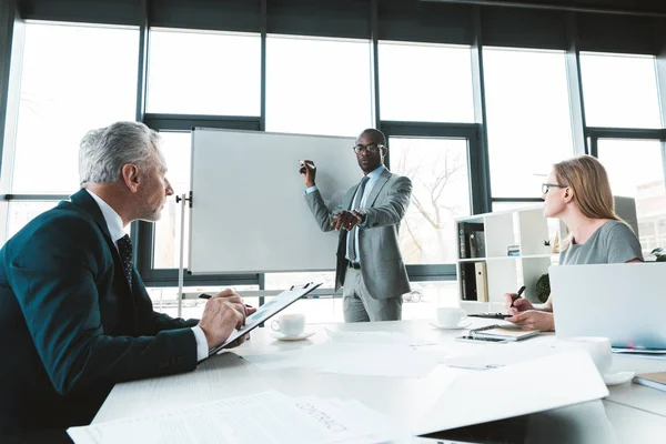 Jeune homme d'affaires afro-américain écrivant sur tableau blanc et regardant ses collègues lors d'une réunion d'affaires — Photo de stock
