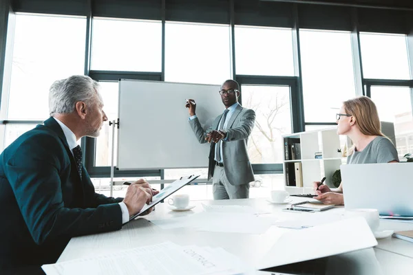 Joven empresario afroamericano escribiendo en pizarra blanca y mirando a sus compañeros de trabajo en la reunión de negocios - foto de stock