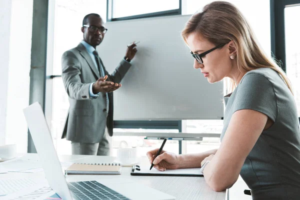 Uomo d'affari afroamericano indicando lavagna bianca e guardando collega donna prendere appunti durante la riunione — Foto stock