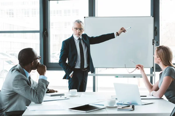 Senior-Geschäftsmann zeigt auf Whiteboard und schaut Kollegen bei Geschäftstreffen an — Stockfoto