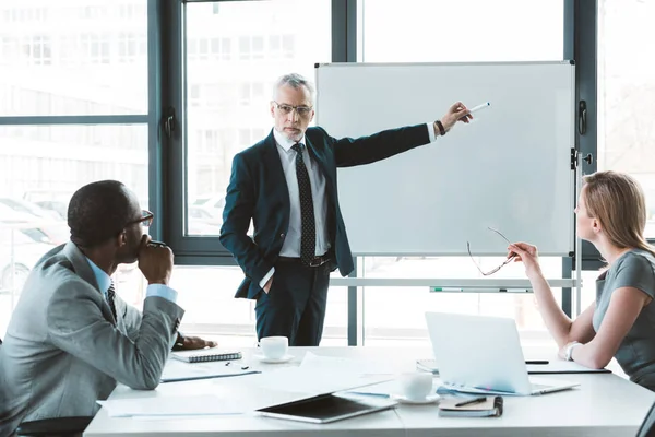 Hombre de negocios profesional senior señalando pizarra blanca y mirando a los colegas durante la reunión de negocios - foto de stock