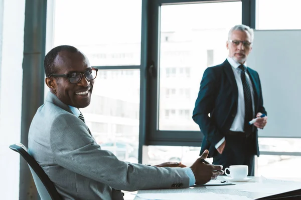 Handsome african american businessman in eyeglasses smiling at camera while working with male colleague in office — Stock Photo
