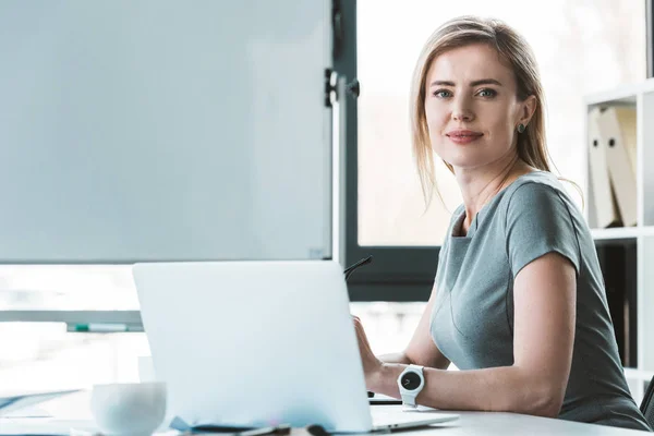 Atractiva mujer de negocios utilizando el ordenador portátil y sonriendo a la cámara - foto de stock