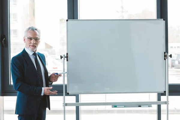 Handsome senior businessman in eyeglasses pointing at blank whiteboard and looking at camera — Stock Photo