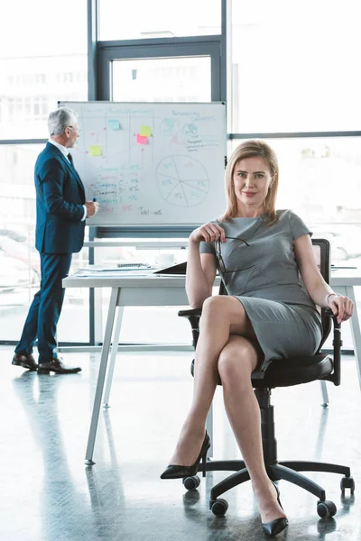 Schöne Geschäftsfrau sitzt auf Stuhl und lächelt in die Kamera, während Senior-Geschäftsmann neben Whiteboard hinter stehen — Stockfoto