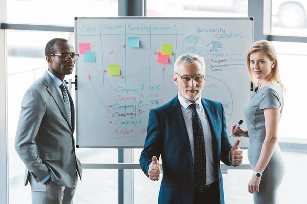 Senior businessman in eyeglasses showing thumbs up and smiling at camera while working with colleagues at whiteboard — Stock Photo