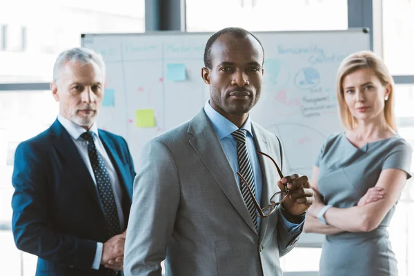 Confiado hombre de negocios afroamericano sosteniendo anteojos y mirando a la cámara mientras colegas de negocios de pie cerca de la pizarra blanca detrás - foto de stock