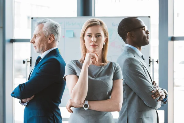 Selbstbewusster Geschäftsmann, der mit der Hand am Kinn in die Kamera blickt, während multiethnische Geschäftsleute am Whiteboard hinter ihm stehen — Stockfoto