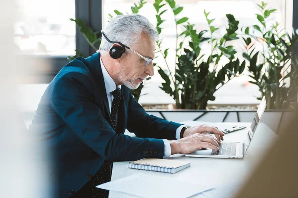 Side view of senior businessman in eyeglasses and headset using laptop in office — Stock Photo