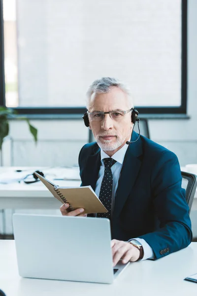 Serious senior businessman in eyeglasses and headset using laptop and looking at camera — Stock Photo