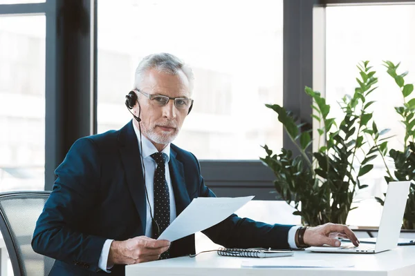 Serious businessman in eyeglasses and headset using laptop and looking at camera — Stock Photo