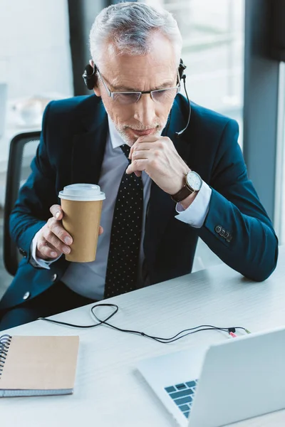 Senior businessman in eyeglasses and headset using laptop and holding paper cup — Stock Photo