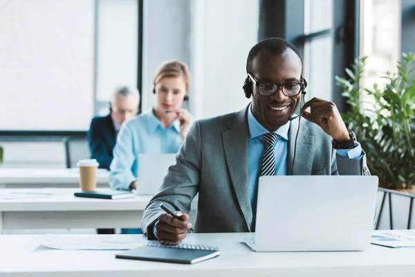 Guapo sonriente afroamericano hombre de negocios en gafas y auriculares usando el ordenador portátil y tomando notas - foto de stock