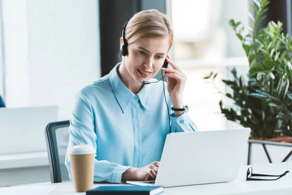 Smiling businesswoman in headset using laptop in office — Stock Photo