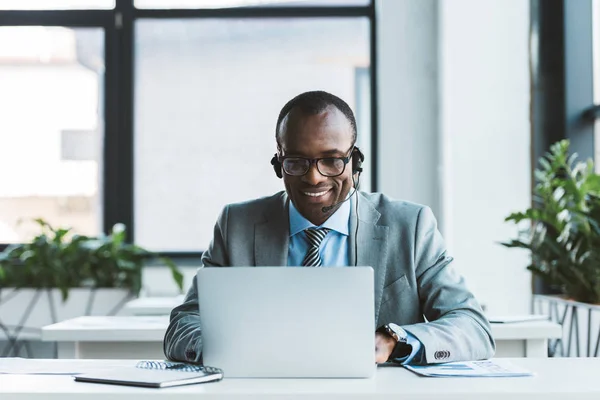 Uomo d'affari africano americano sorridente in occhiali e auricolare utilizzando il computer portatile in ufficio — Foto stock