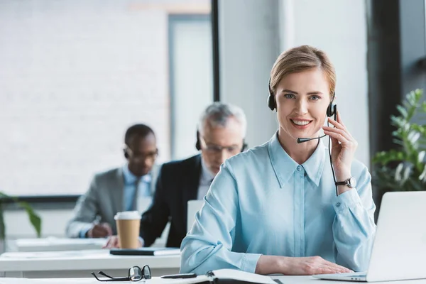 Attraente donna d'affari in auricolare utilizzando il computer portatile e sorridente alla fotocamera — Foto stock