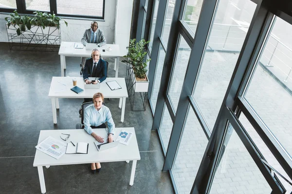 High angle view of multiethnic business people sitting at tables and looking at camera in modern office — Stock Photo