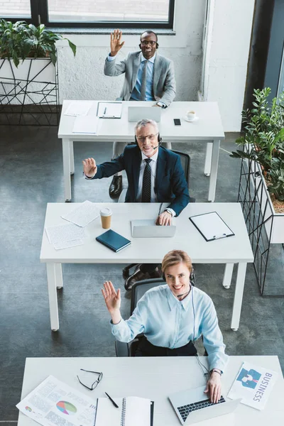 High angle view of cheerful business people in headsets smiling at camera and waving hands — Stock Photo