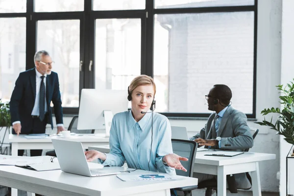 Empresária em fone de ouvido usando laptop e olhando para a câmera enquanto colegas do sexo masculino que trabalham para trás no escritório — Fotografia de Stock