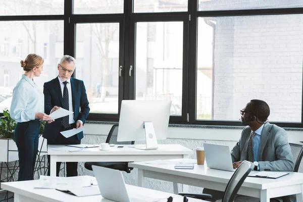 Hombre de negocios afroamericano utilizando el ordenador portátil y mirando a los colegas discutiendo documentos en la oficina - foto de stock